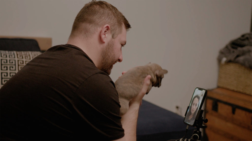 Peter K doing a facetime interview with a customers. Peter Is sitting down wearing a black t-shirt holding a brindle puppy in his right hand .