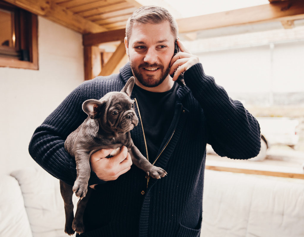 Peter K holding Füli as a puppy. Füli is described a Blue French Bulldog. Peter is a wearing a knitted blue sweater with a gold zipper and a black tee shirt. He is holding up a phone to his left ear. 
